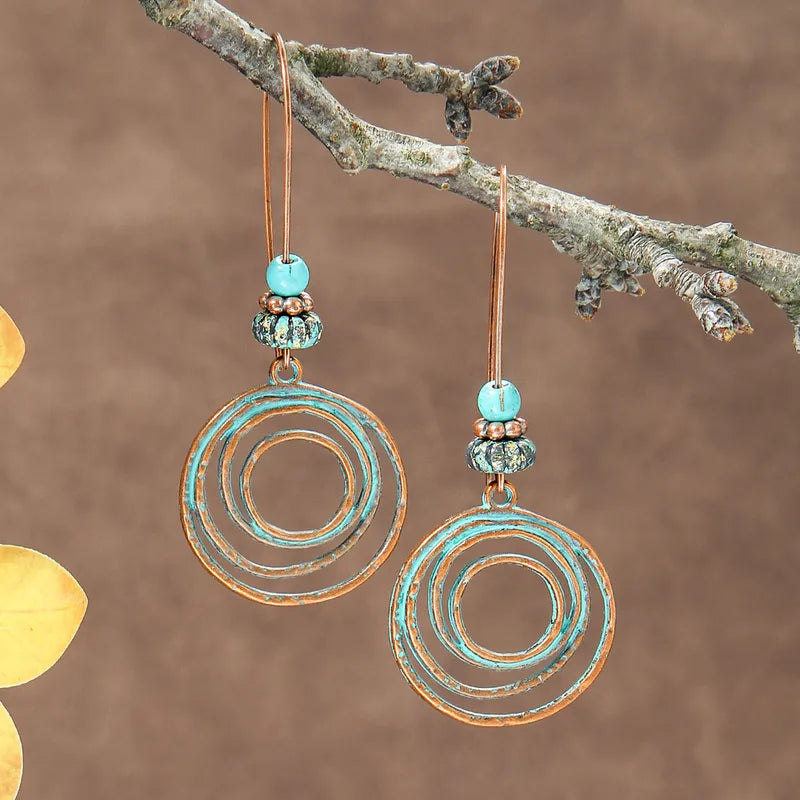 A pair of oxidised boho earrings with turquoise beads and concentric patina circles, hanging from a tree branch.