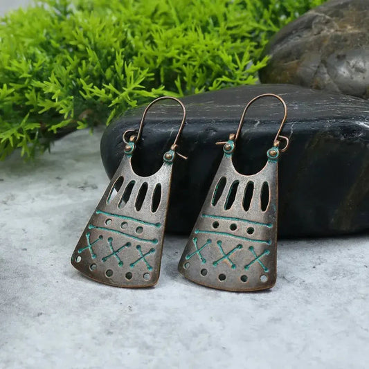 A pair of oxidised earrings with a tribal geometric design in bronze and teal, set against a grey background with green foliage and dark rocks.