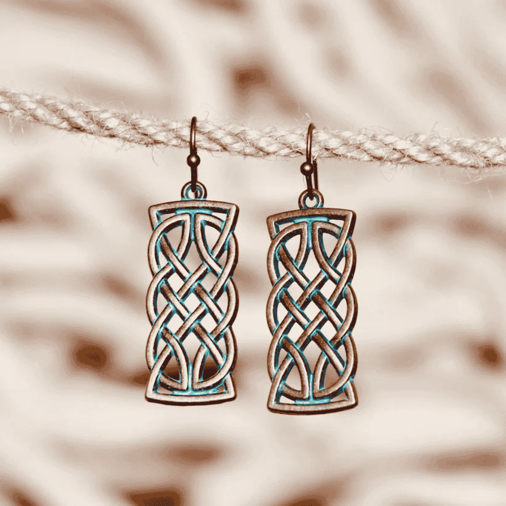 A pair of oxidised earrings with a Celtic knot design and blue patina, hanging on a rope against a blurred background.