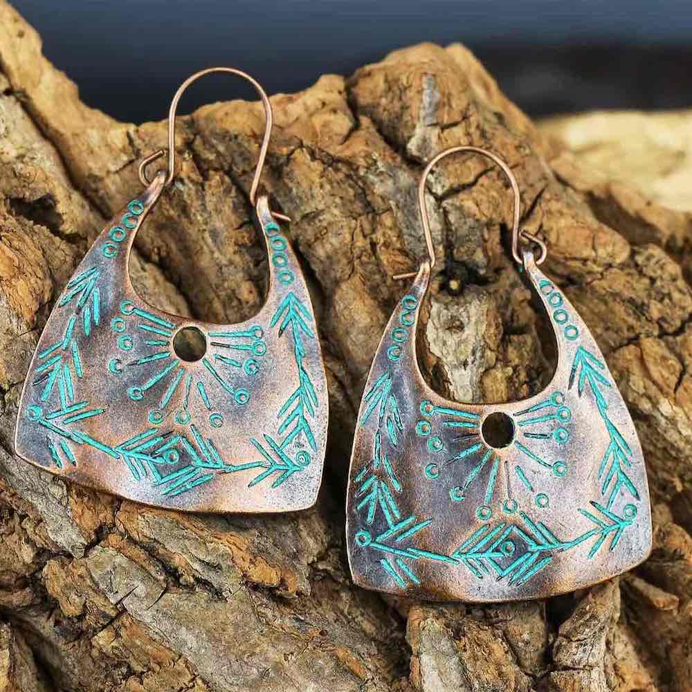 A pair of oxidised copper boho drop earrings with turquoise patina details, displayed on a piece of rustic wood.