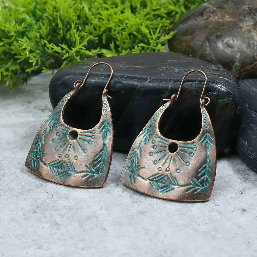 Two oxidised copper boho drop earrings with turquoise accents, resting on a dark stone with green foliage in the background.