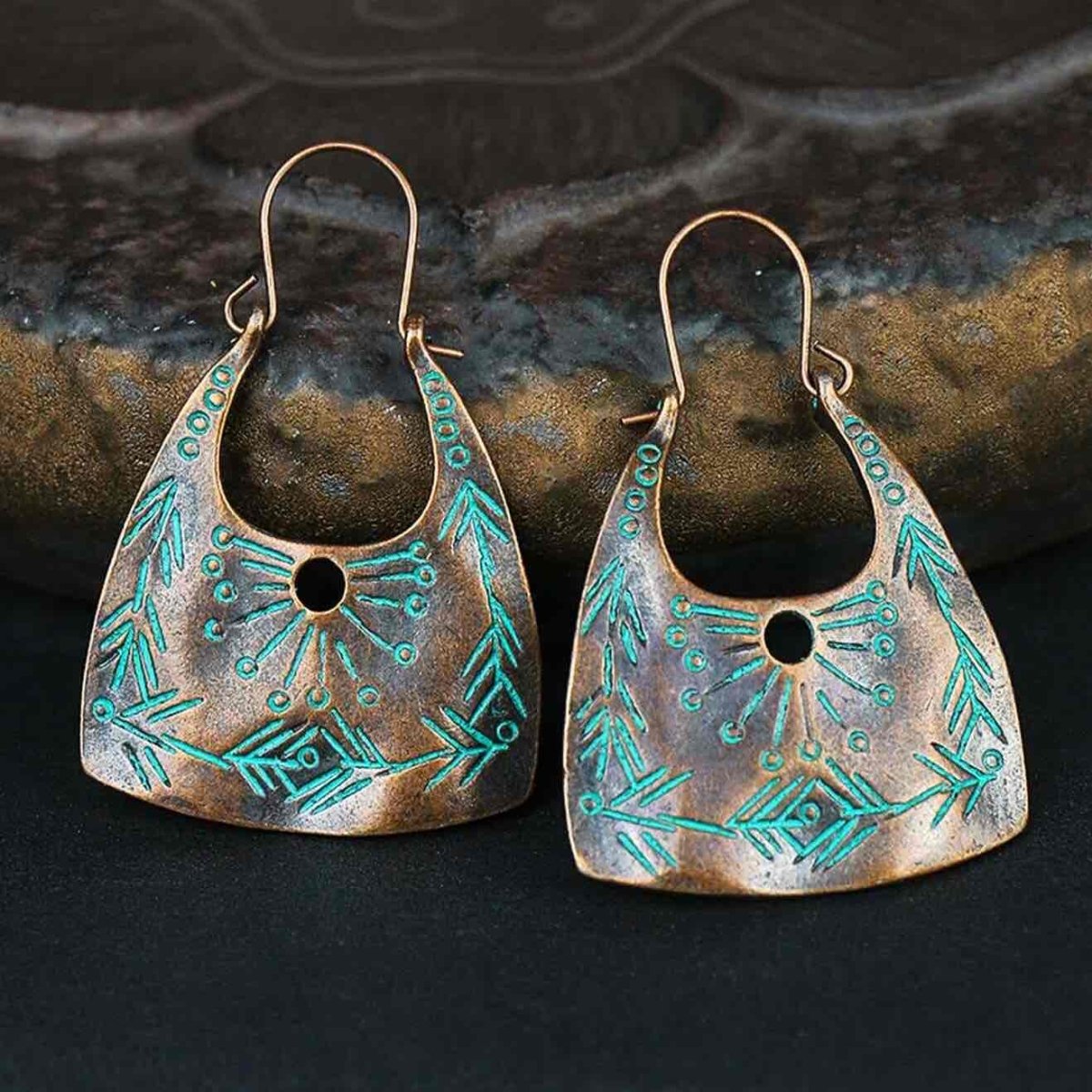 A close-up of two oxidised copper boho drop earrings featuring turquoise patterns, set against a dark, textured background.