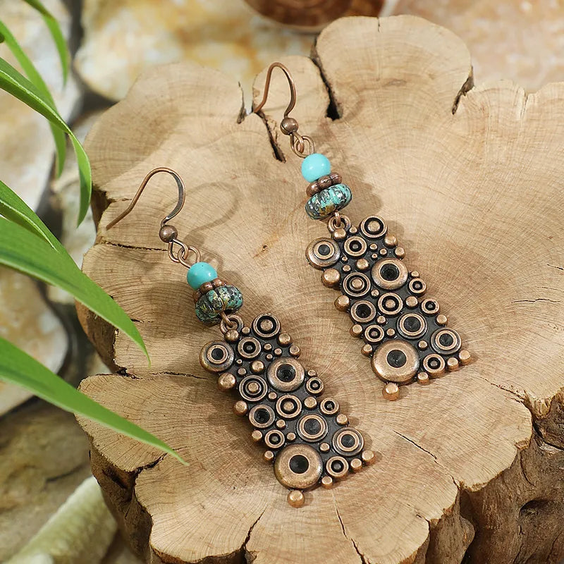 Pair of oxidised copper dangle earrings with concentric circles and turquoise beads, resting on a rustic wooden slab.