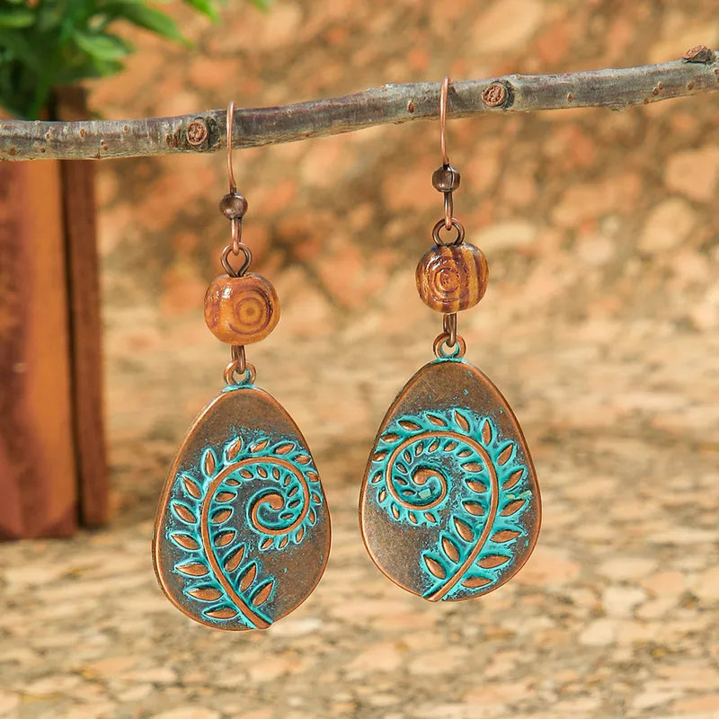 A pair of oxidised earrings with a copper base and teal leaf swirl design, hanging from a branch.