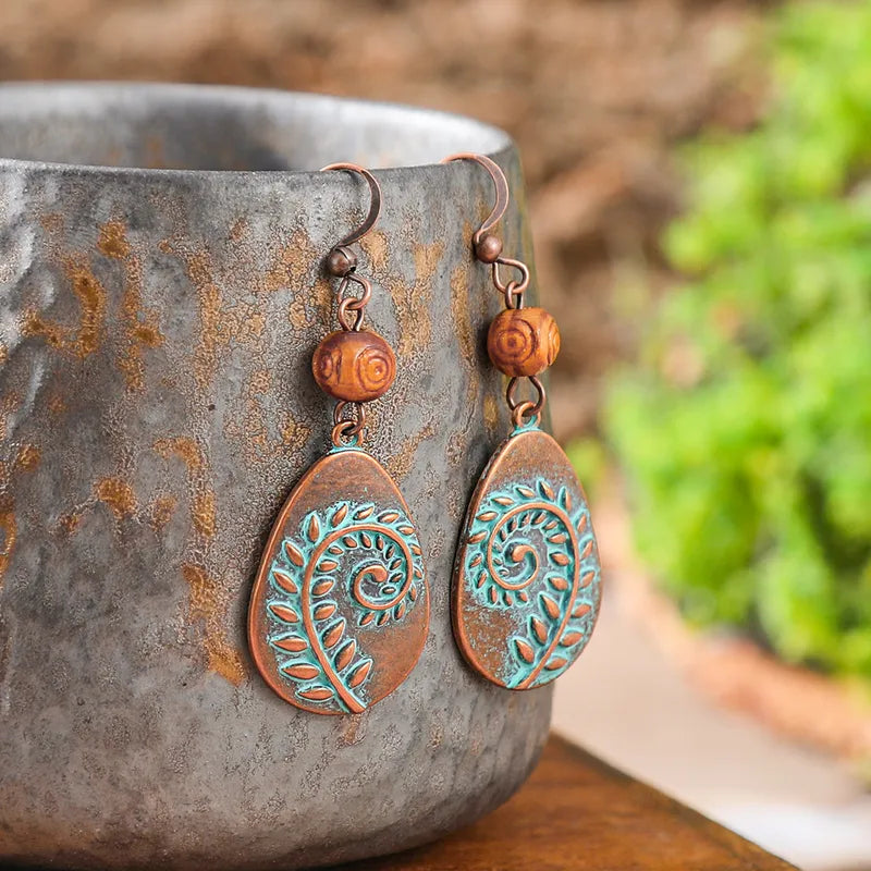 A pair of oxidised earrings with a copper base and teal leaf swirl design, resting on a textured grey pot.
