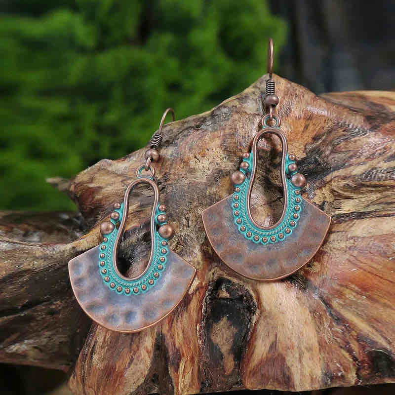 A pair of oxidised earrings with a fan shape and teal patina, displayed on a wooden surface with green foliage in the background.