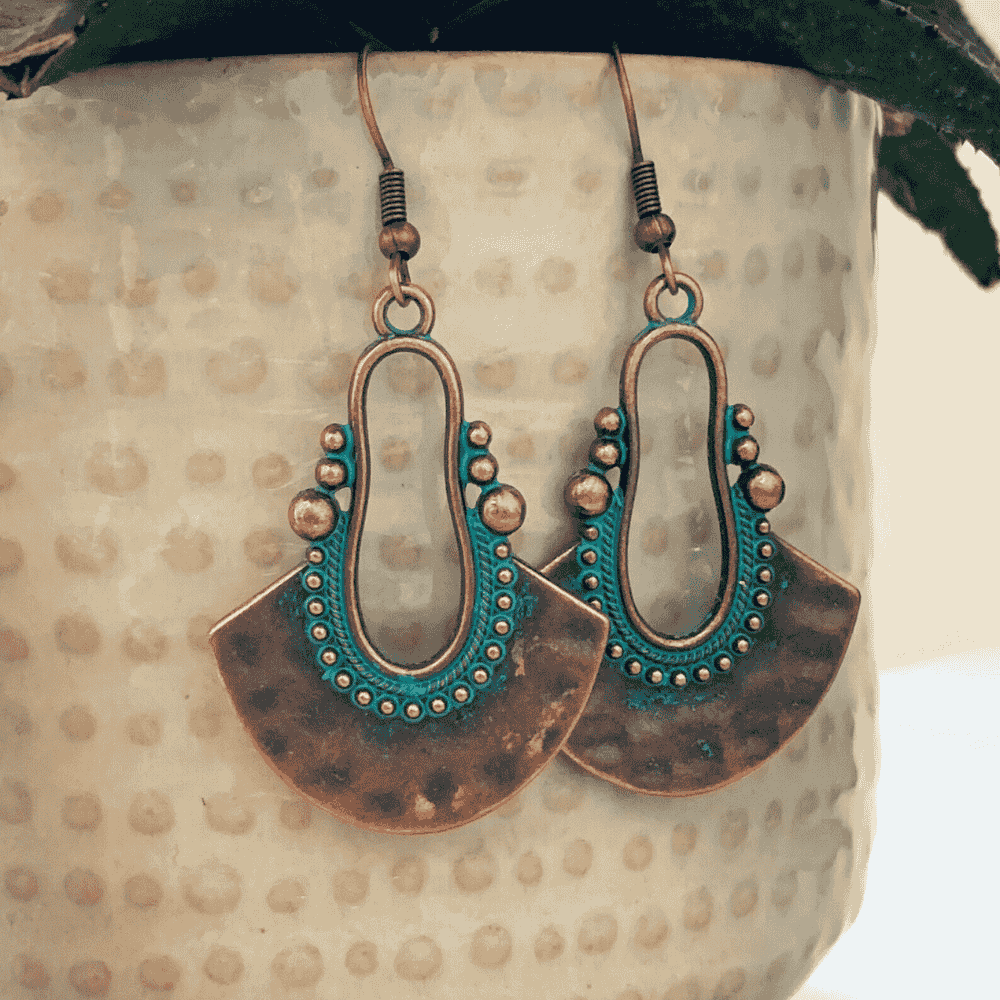 A pair of oxidised earrings with a fan shape and teal patina, hanging against a textured white pot with a plant in the background.