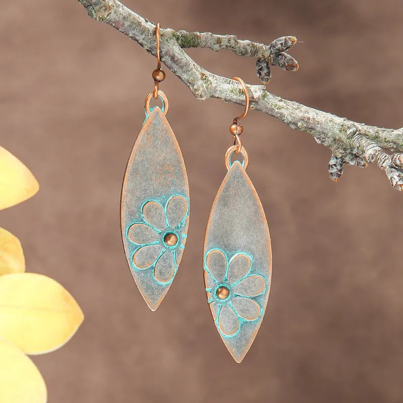 A pair of oxidised earrings with a floral teal patina design, hanging from a tree branch, highlighting their long dangle style.