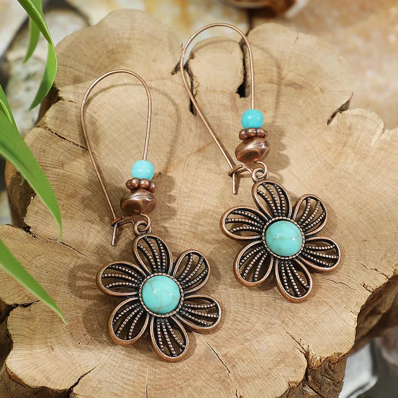 A pair of oxidised earrings with copper floral drops and turquoise stones, set against a rustic wooden background.