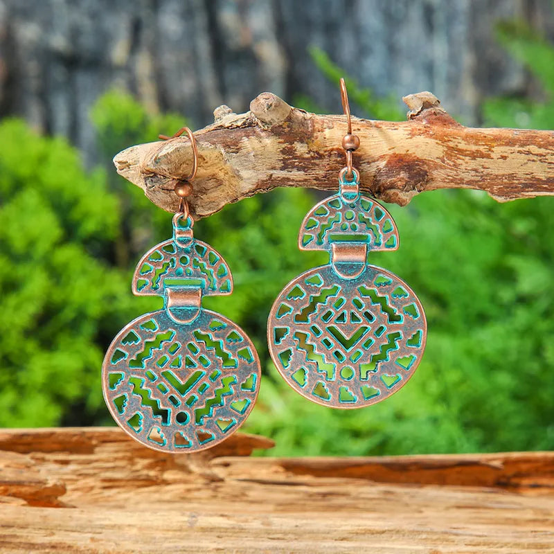 A pair of oxidised earrings with a geometric pattern and patina finish, hanging from a wooden branch against a blurred green background.