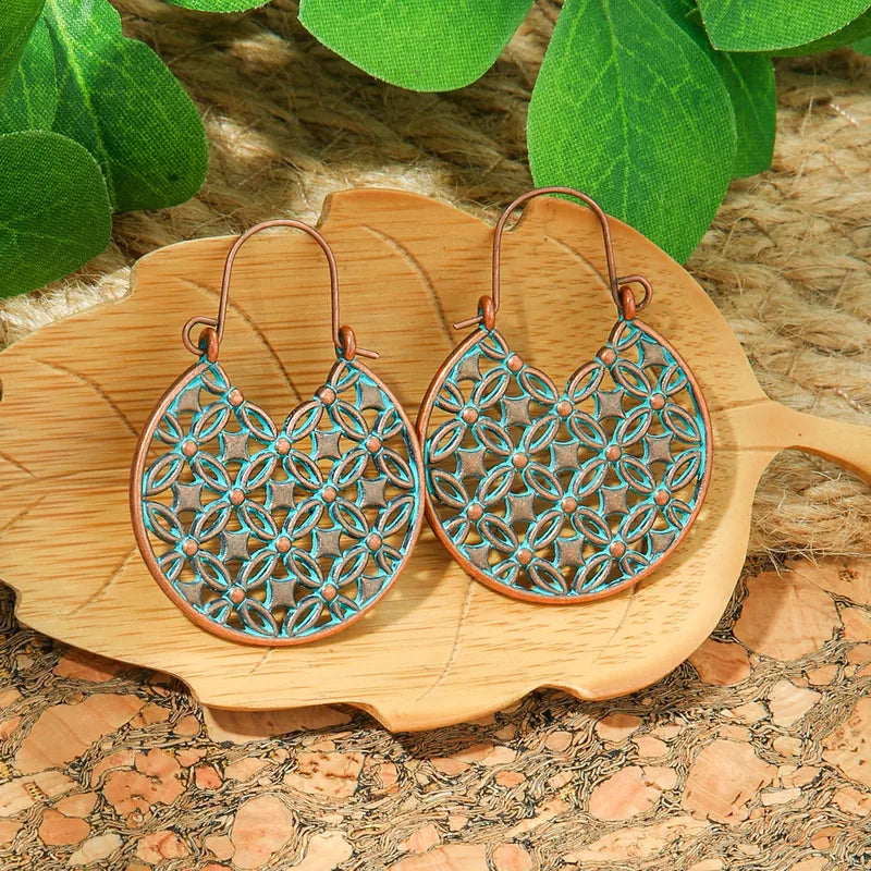 A pair of oxidised earrings with a teardrop shape and intricate geometric pattern in copper and teal, displayed on a wooden leaf-shaped plate.