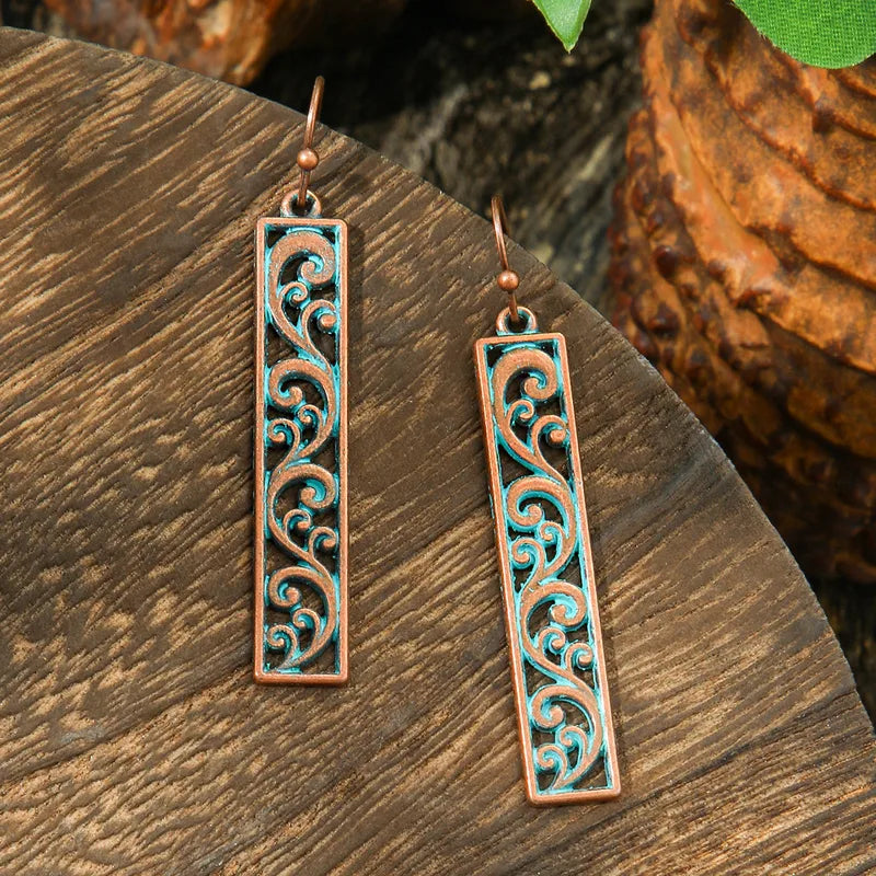 A pair of oxidised earrings with a rectangular bar shape, featuring intricate teal filigree scrollwork on a copper-toned base, displayed on a wooden surface.