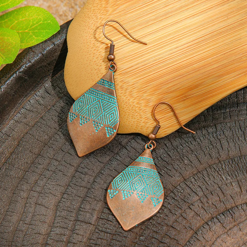 A pair of oxidised earrings with copper and turquoise blue geometric patterns, lying on a dark textured surface next to a wooden block.