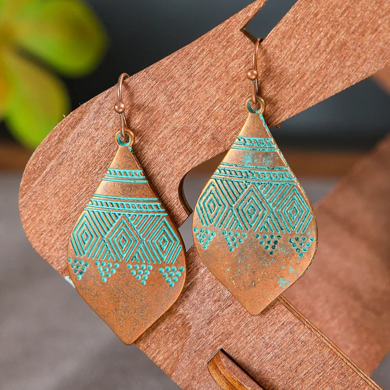 Close-up of a pair of oxidised earrings, copper with turquoise blue geometric designs, displayed on a light brown wooden stand.