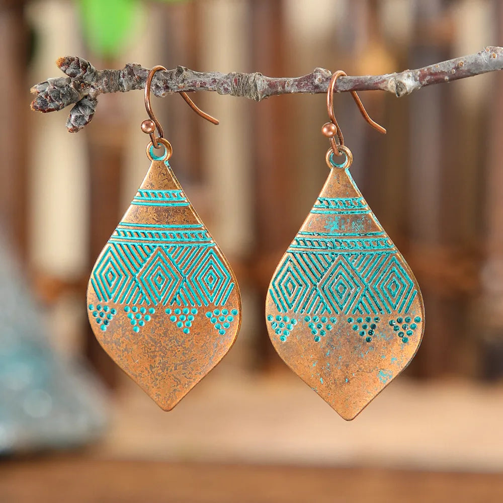 A pair of oxidised earrings, copper with turquoise blue geometric patterns, hanging from a small tree branch against a blurred background.
