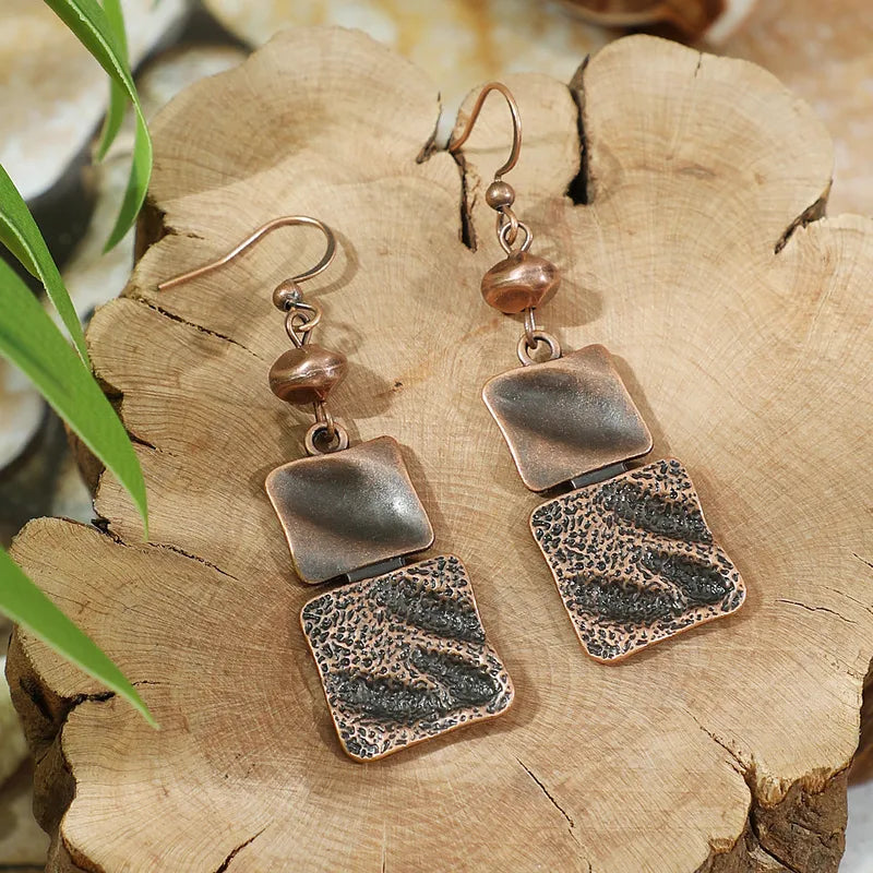 A pair of oxidised copper textured dangle earrings with square elements, resting on a wooden surface, showcasing their rustic design.