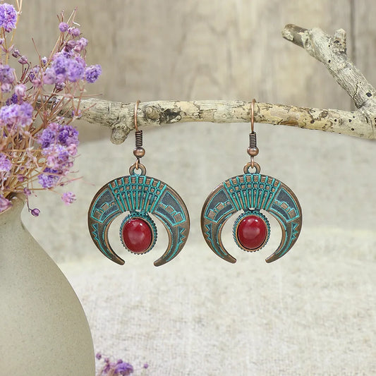A pair of oxidised crescent drop earrings with a red stone and blue patina finish, hanging from a branch next to a vase of purple flowers.