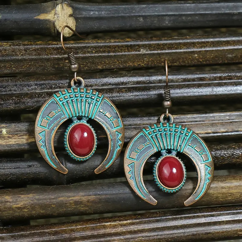 Close-up of a pair of oxidised crescent drop earrings featuring a red stone and intricate blue patina detailing, set against a dark wooden background.