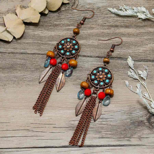 A pair of oxidised dreamcatcher long earrings with turquoise and red beads, and bronze chains, laid on a wooden surface.