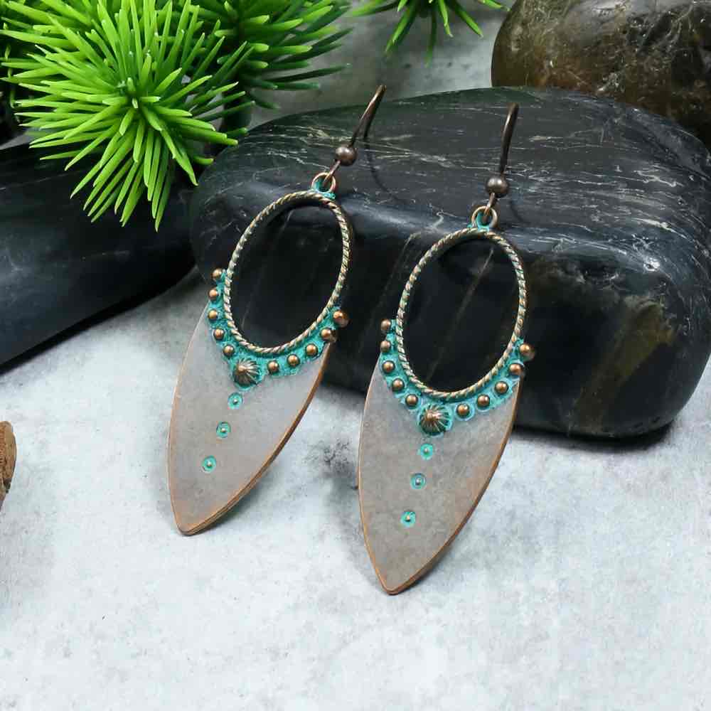 A pair of oxidised earrings with teal accents and a dual-tone finish, displayed on a grey surface with green foliage and dark stones in the background.