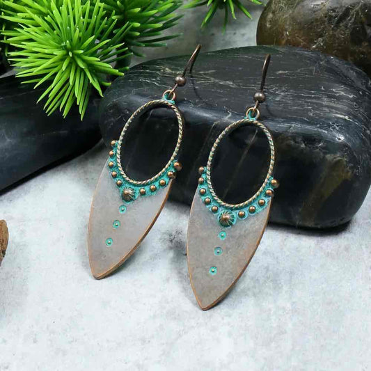 A pair of oxidised earrings with teal accents and a dual-tone finish, displayed on a grey surface with green foliage and dark stones in the background.