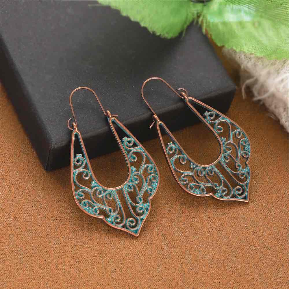 A pair of oxidised filigree drop earrings, featuring a patina green design, placed on a brown surface with a black box in the background.