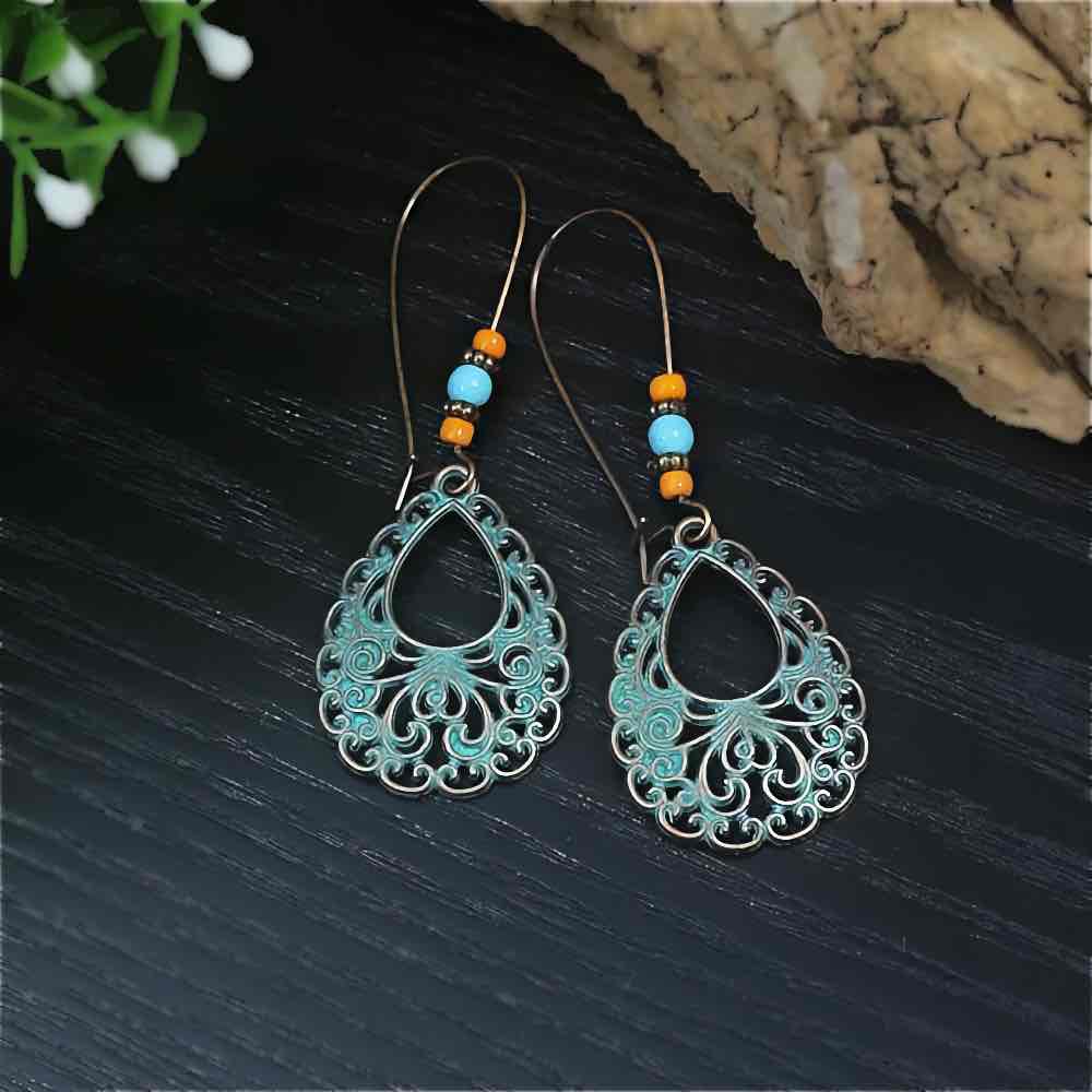 A pair of oxidised earrings with teal filigree teardrops and orange and turquoise beads, displayed on a dark wooden surface.