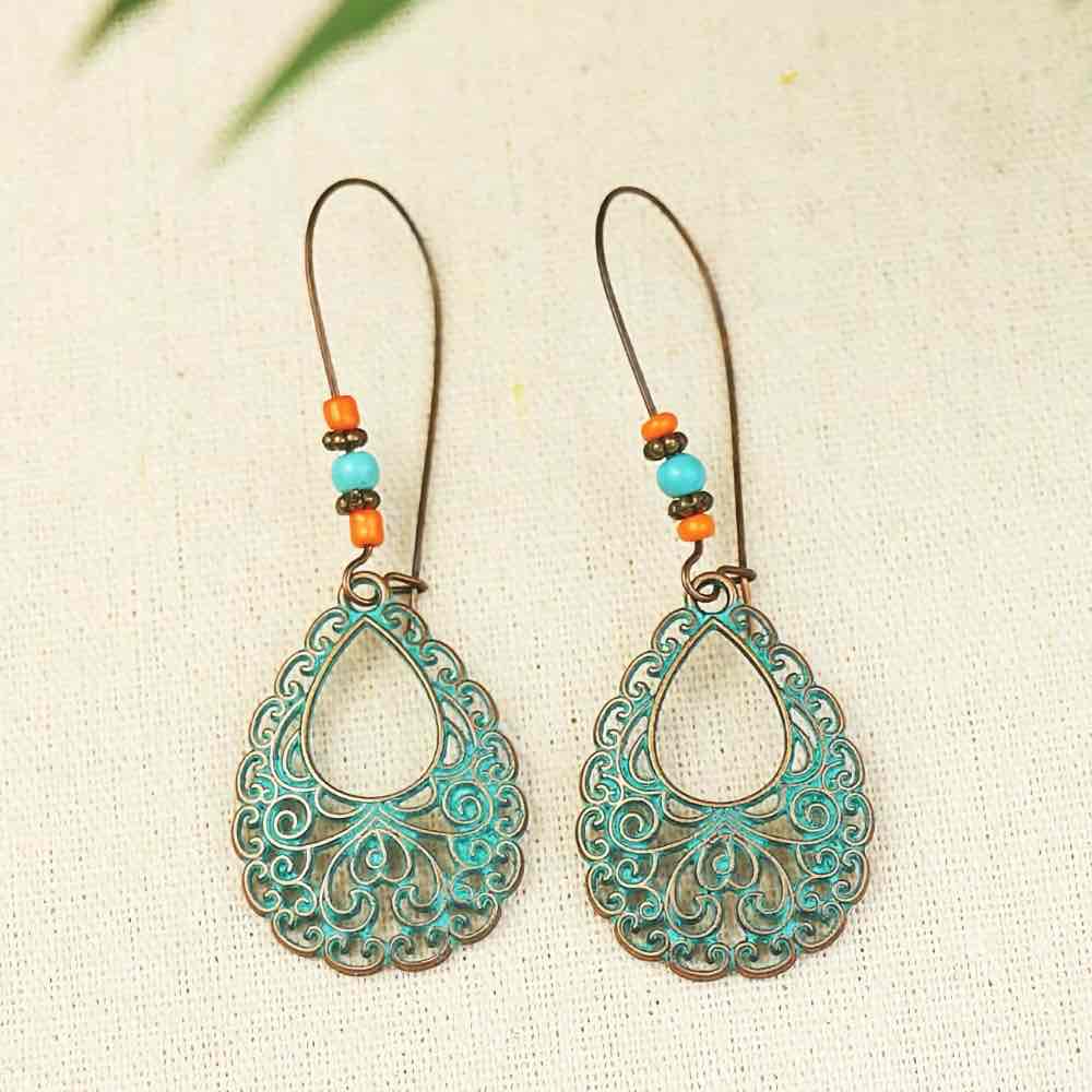 A pair of oxidised earrings with teal filigree teardrops and orange and turquoise beads, shown on a light fabric background.