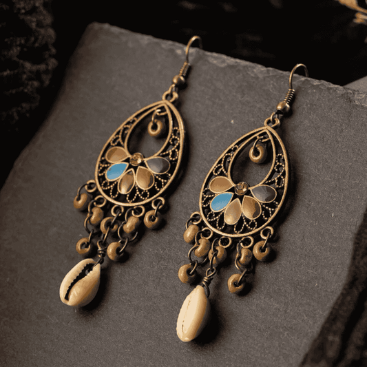 A pair of oxidised floral shell drop earrings with blue and black enamel details, resting on a dark textured surface.