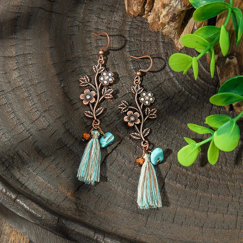 A pair of oxidised floral tassel drop earrings with turquoise accents, displayed on a dark wooden surface with green leaves in the background.