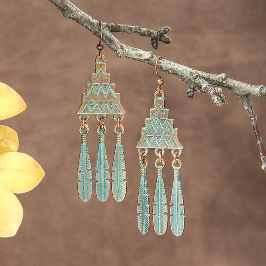 Close-up of oxidised geometric feather dangle earrings with teal patina, hanging from a tree branch.