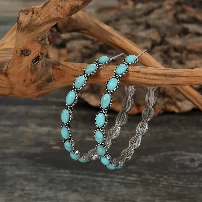 A pair of oxidised silver hoop earrings with oval turquoise stones and a twisted back, displayed on a natural wooden branch.