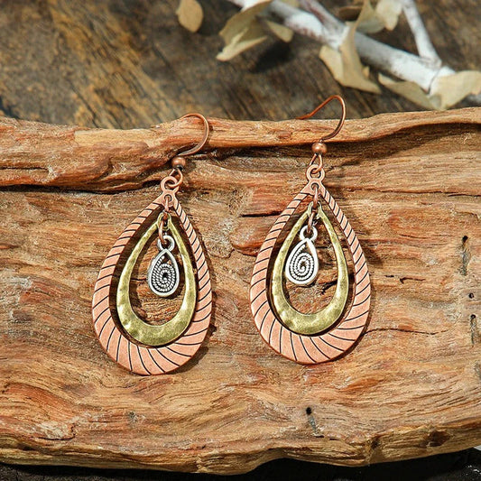 A pair of oxidised earrings with layered teardrop shapes in copper, antique gold, and silver, displayed on a wooden surface.