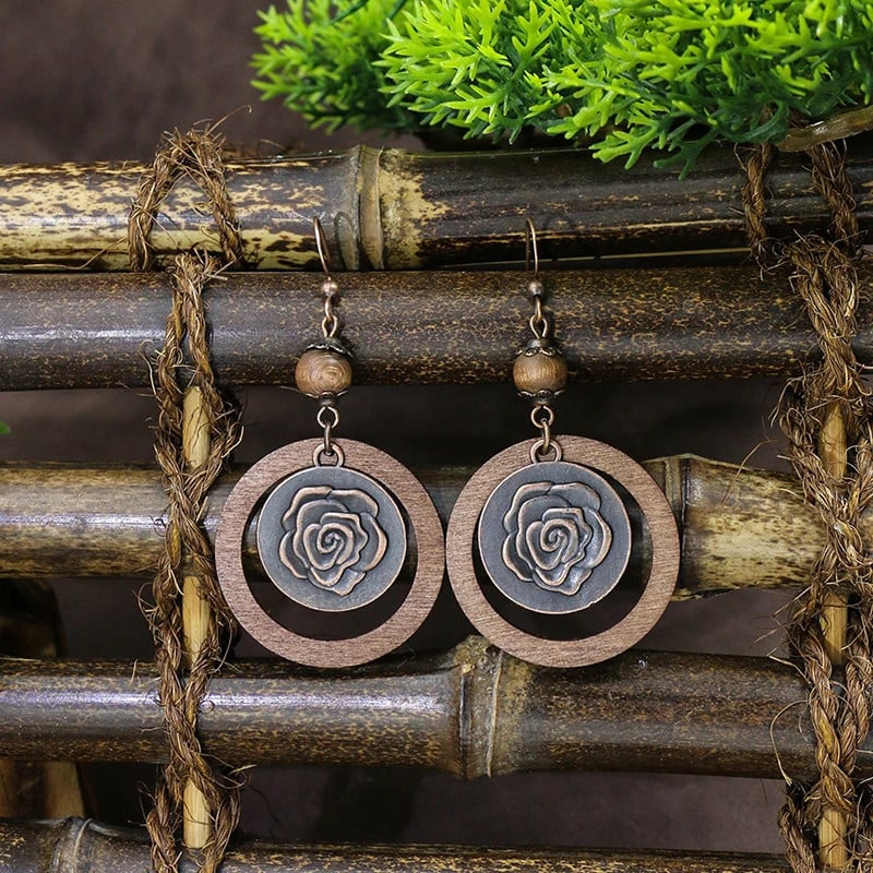 Pair of oxidised earrings with a rose design and wooden hoops, hanging on bamboo sticks with green foliage in the background.
