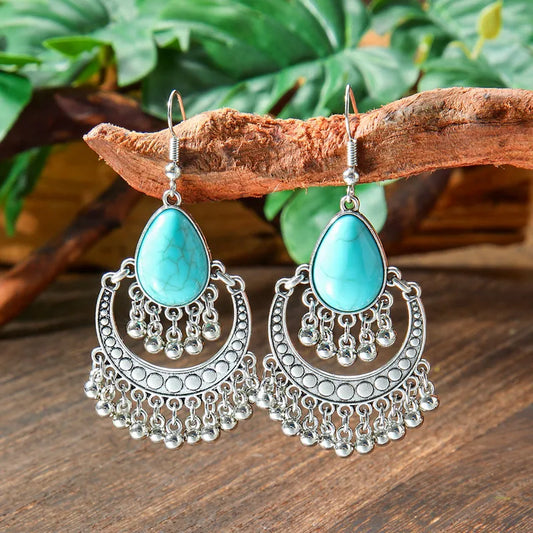 A pair of oxidised silver drop earrings with teardrop turquoise stones hanging from a wooden branch, with people in the blurred background.