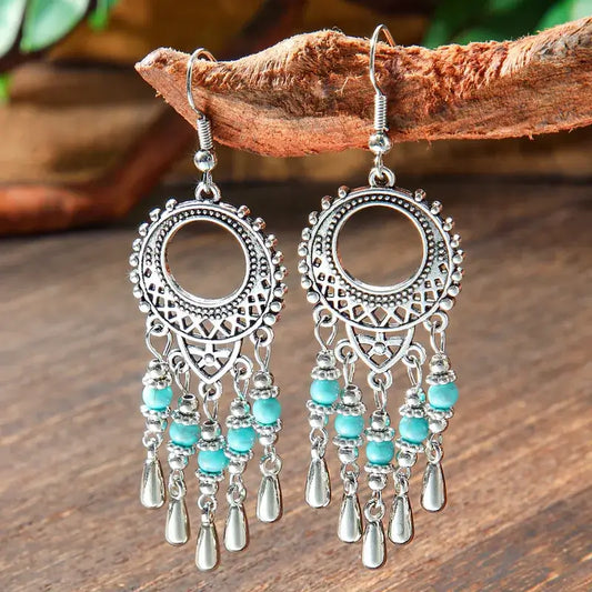 A pair of oxidised earrings with circular filigree and dangling turquoise beads, hanging from a rustic wooden branch.