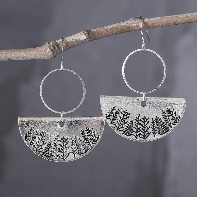 A pair of oxidised earrings with a forest fern design hanging from a tree branch.
