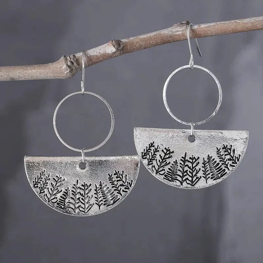 A pair of oxidised earrings with a forest fern design hanging from a tree branch.