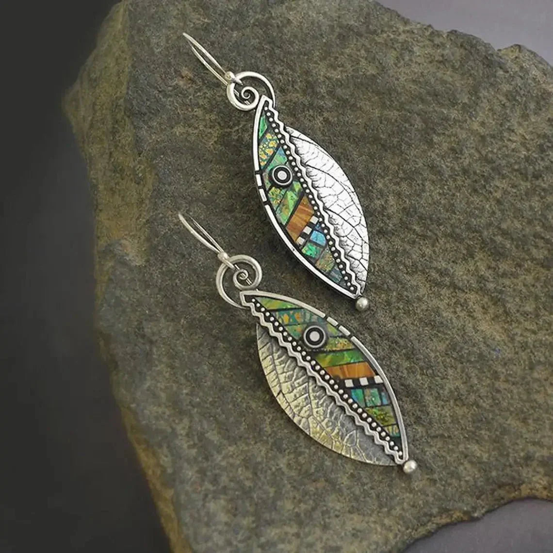 Two oxidised earrings, designed as textured leaves with colorful mosaic and filigree details, resting on a natural stone surface.