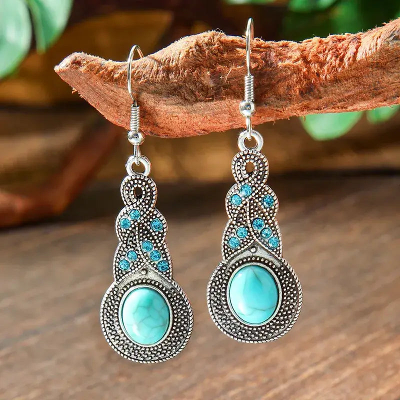 A pair of oxidised earrings with turquoise oval stones and intricate silver detailing, hanging from a tree branch in a natural setting.