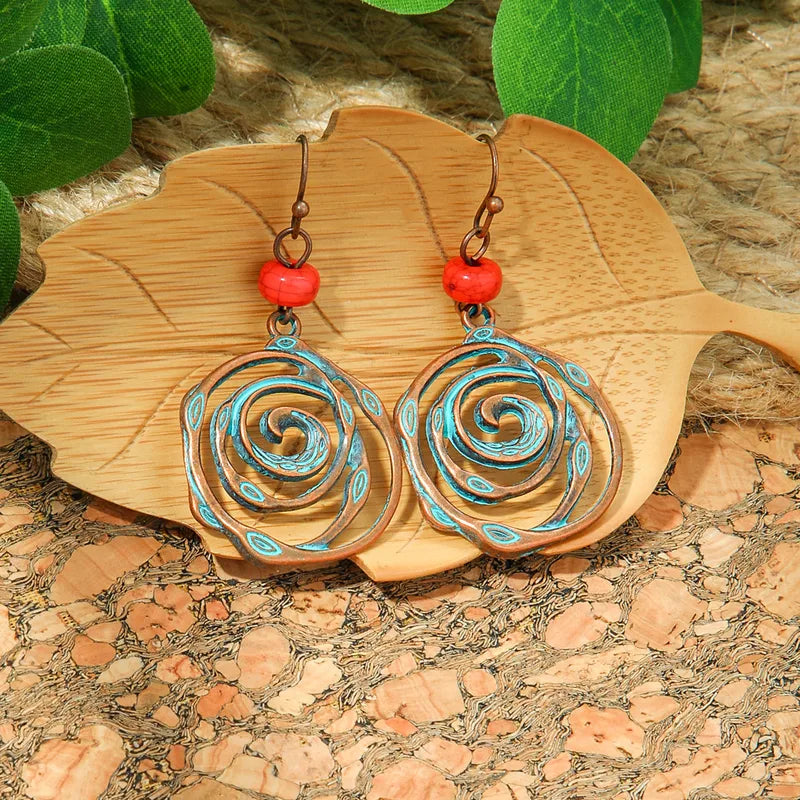 A pair of oxidised spiral dangle earrings with blue patina and a red bead, resting on a light wooden leaf-shaped tray against a textured brown background with green leaves.