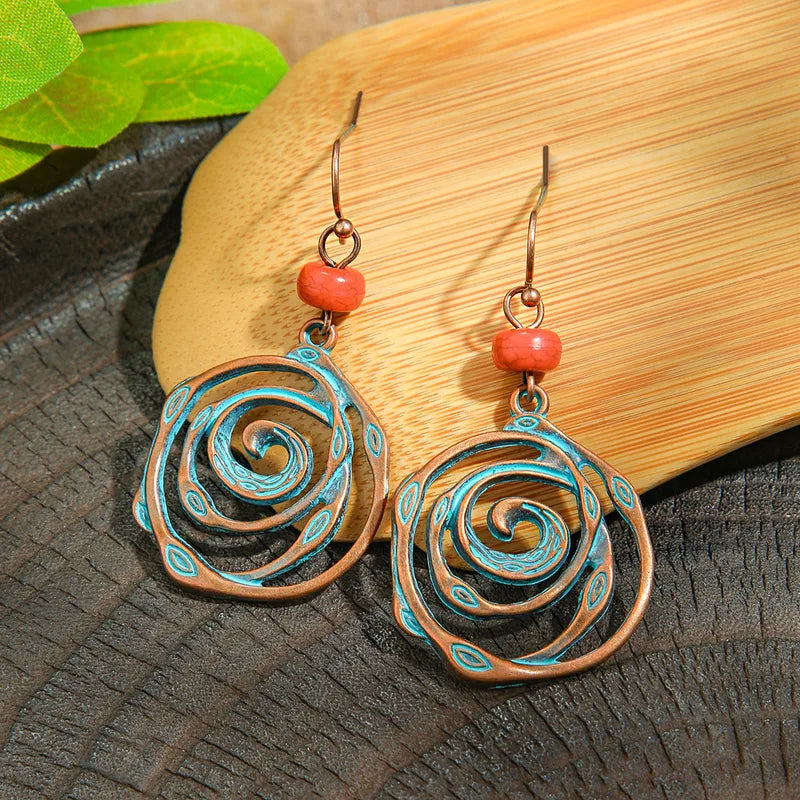 A pair of oxidised spiral dangle earrings with blue patina and a red bead, displayed on a dark wooden surface with a light wooden leaf-shaped tray and green leaves in the background.