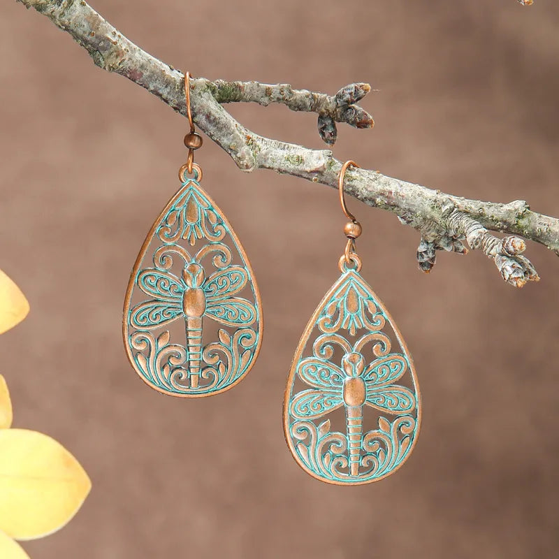 A pair of oxidised earrings with a teal dragonfly filigree design, hanging from a tree branch.