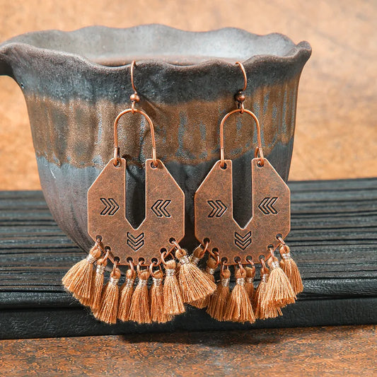 A pair of oxidised tribal geometric tassel drop earrings with a copper finish, hanging against a dark ceramic pot.