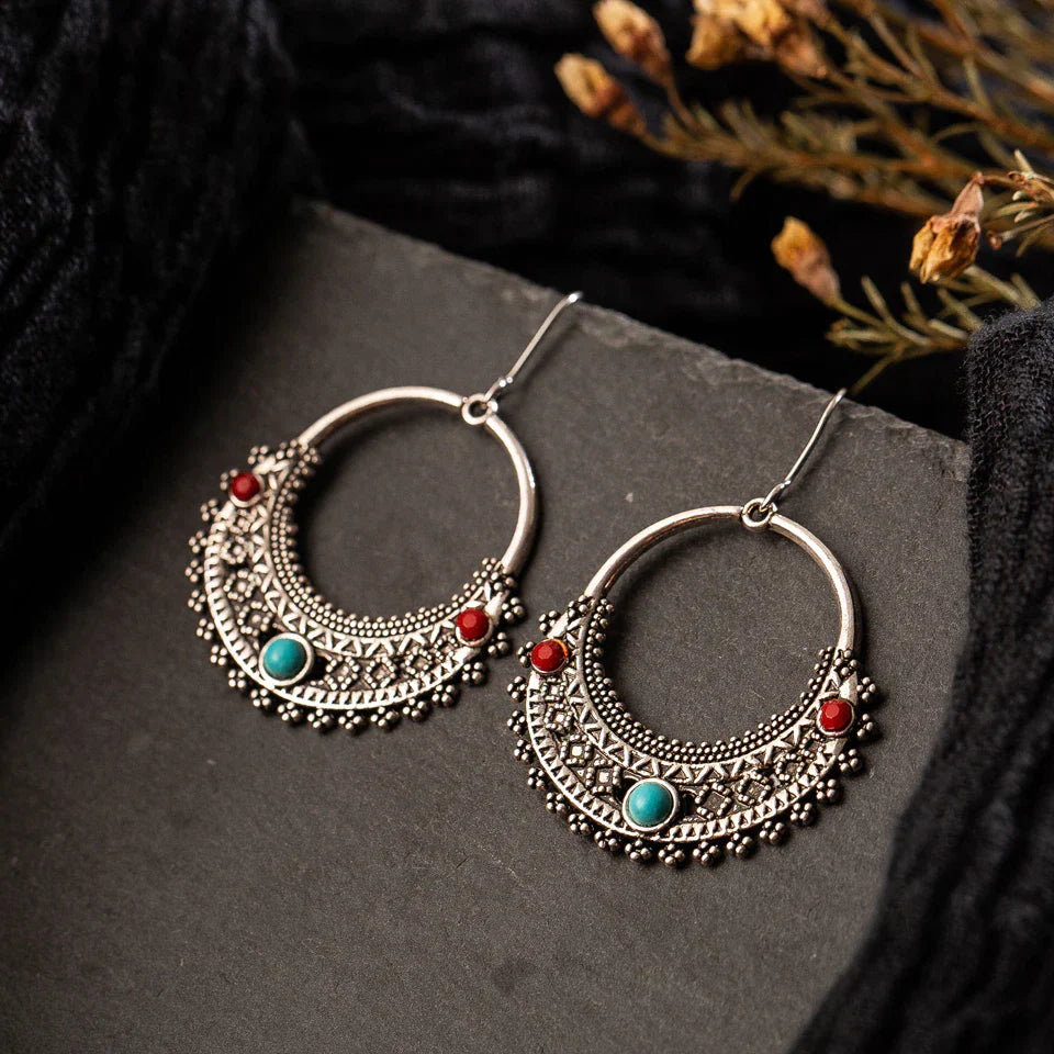A pair of oxidised tribal hoop earrings with red and turquoise stones displayed on a dark surface, highlighting their intricate design. These oxidised earrings are perfect for a bohemian statement.