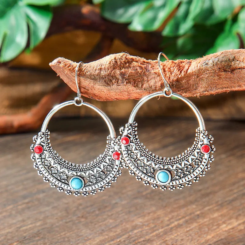 Two oxidised tribal hoop earrings with red and turquoise stones hanging from a tree branch, showcasing their bohemian charm. These oxidised earrings are a beautiful accessory.