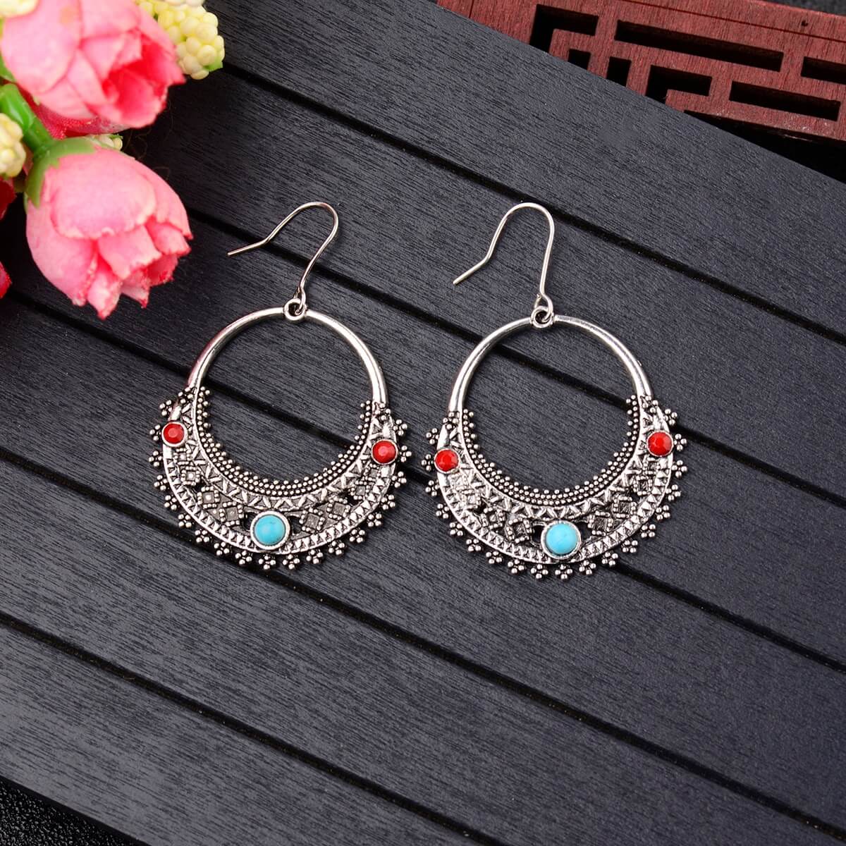 A pair of oxidised tribal hoop earrings with red and turquoise stones laid on a black wooden surface, with pink flowers in the background. These oxidised earrings feature a unique design.