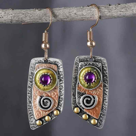 Close-up of a pair of oxidised earrings with purple stones and tribal spiral designs, hanging from a branch.