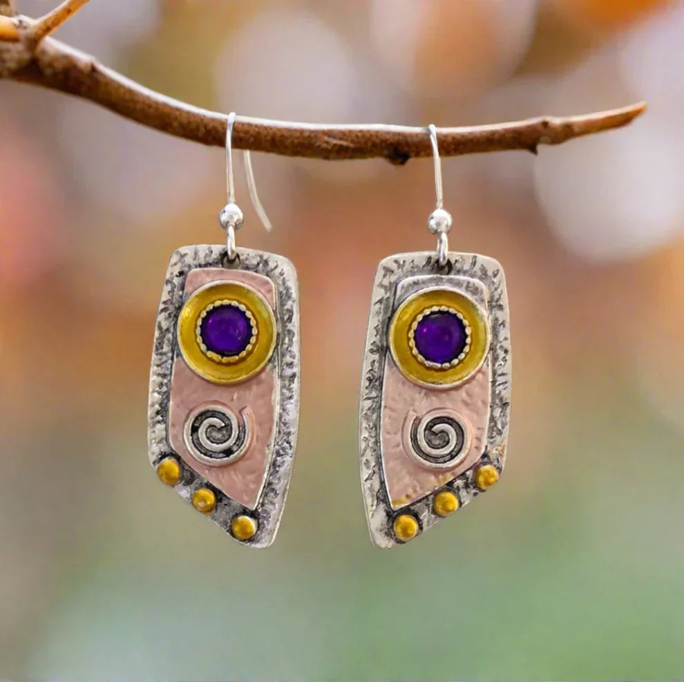 A pair of oxidised earrings with purple stones and tribal spiral designs, hanging from a branch against a blurred background.