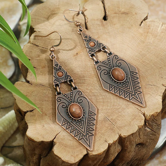 A pair of oxidised tribal stone drop earrings with bohemian dangle design, featuring brown stones and geometric patterns, displayed on a wooden surface with green leaves.